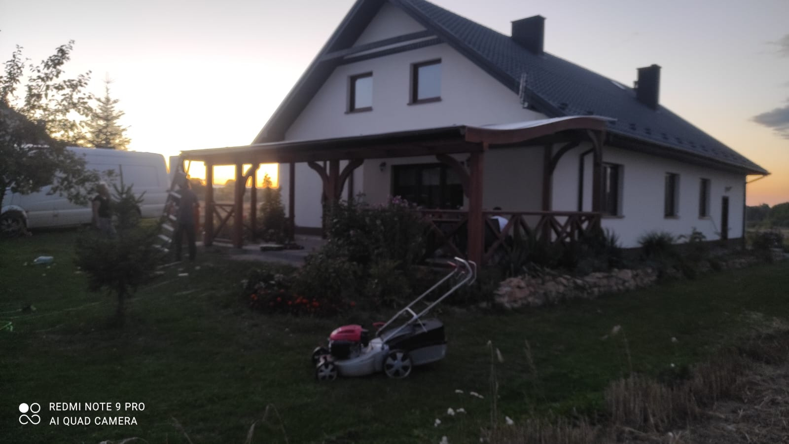 Dom jednorodzinny z drewnianą konstrukcją tarasu w Łazach dużych, widok z trawnika, w tle sylwetki dwóch osób i bus.