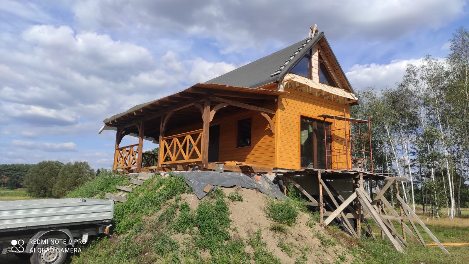 Żółty dom w trakcie budowy z drewnianą werandą, widoczny z boku na wzniesieniu, z elementami rusztowania i przyczepą w tle.
