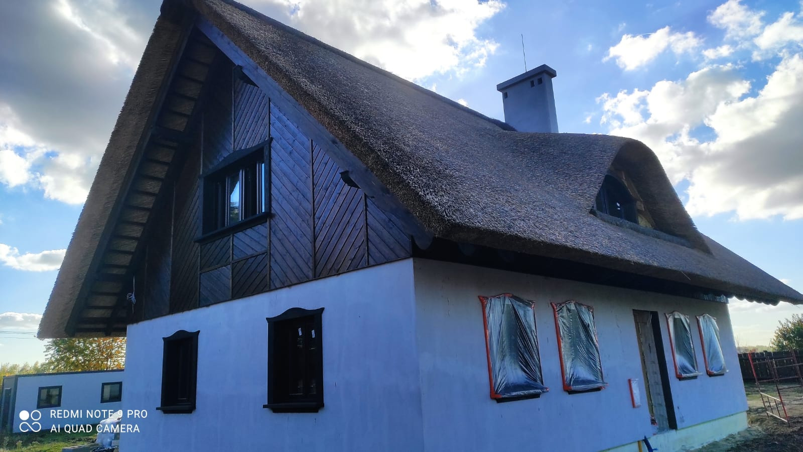 Dom z dachem krytym strzechą, okna zabezpieczone folią, drewniana elewacja szczytowa z ukośnymi deskami, widoczny komin i fragment nieba z chmurami.