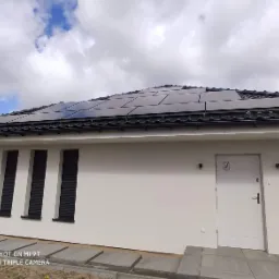 Panele słoneczne na dachu nowoczesnego, parterowego domu z trzema oknami z żaluzjami zewnętrznymi i białymi ścianami, widoczne betonowe płyty chodnikowe i fragment pochmurnego nieba.