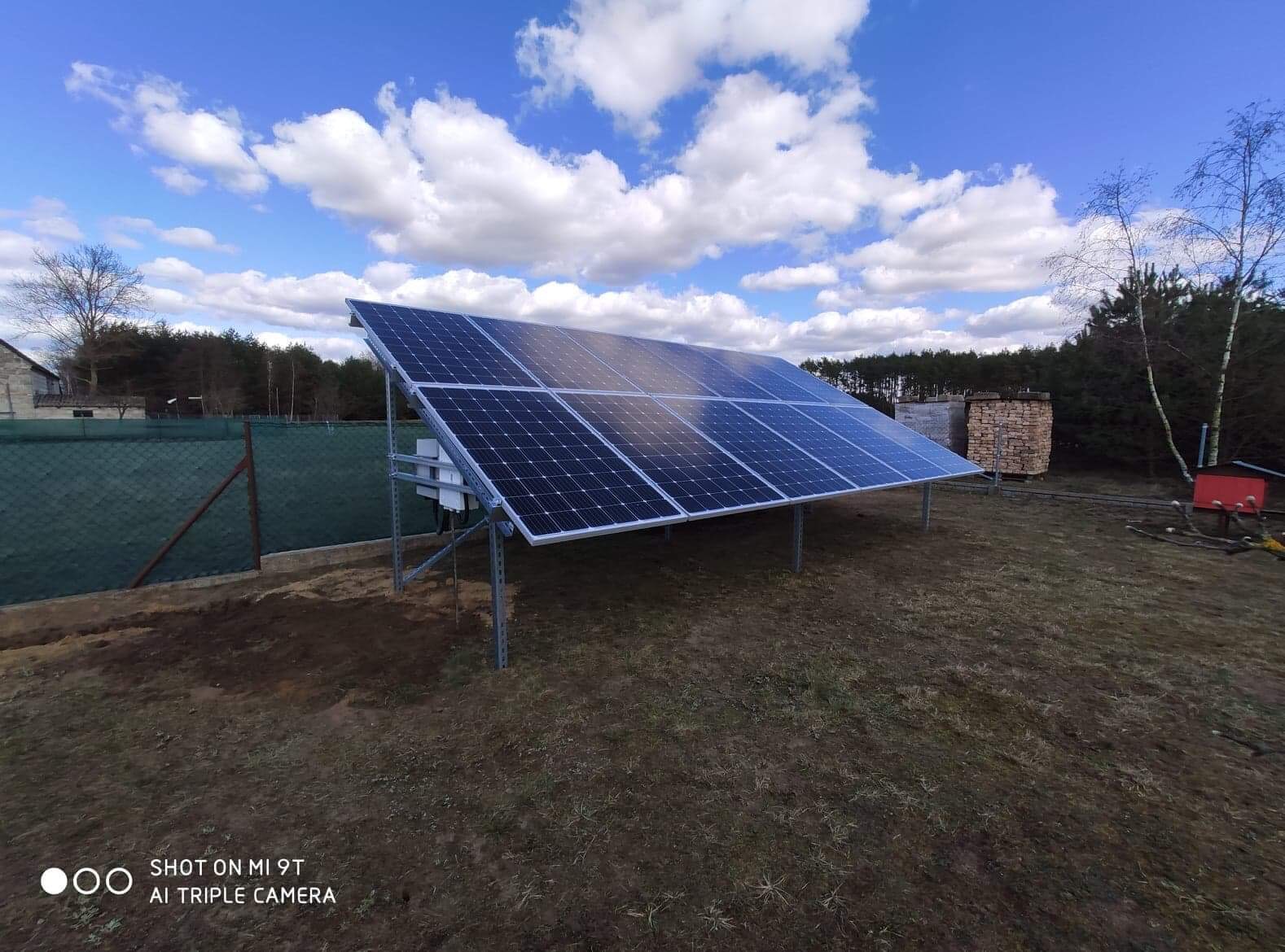 Zestaw paneli słonecznych na metalowej konstrukcji, zainstalowany na trawiastym terenie z widocznym ogrodzeniem i budynkiem w tle, sfotografowany pod błękitnym niebem z chmurami.