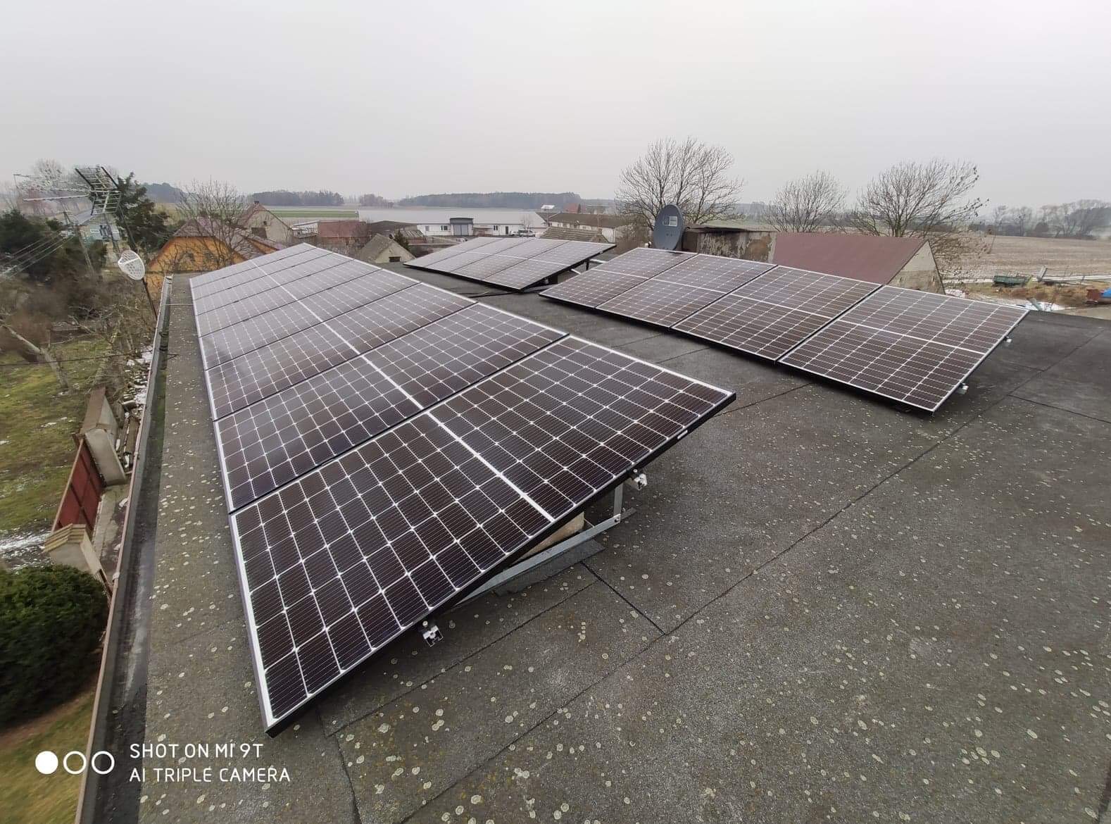 Instalacja fotowoltaiczna na płaskim dachu pokrytym papą, widok z góry, w tle zabudowania wiejskie i zbiornik wodny w pochmurny dzień.
