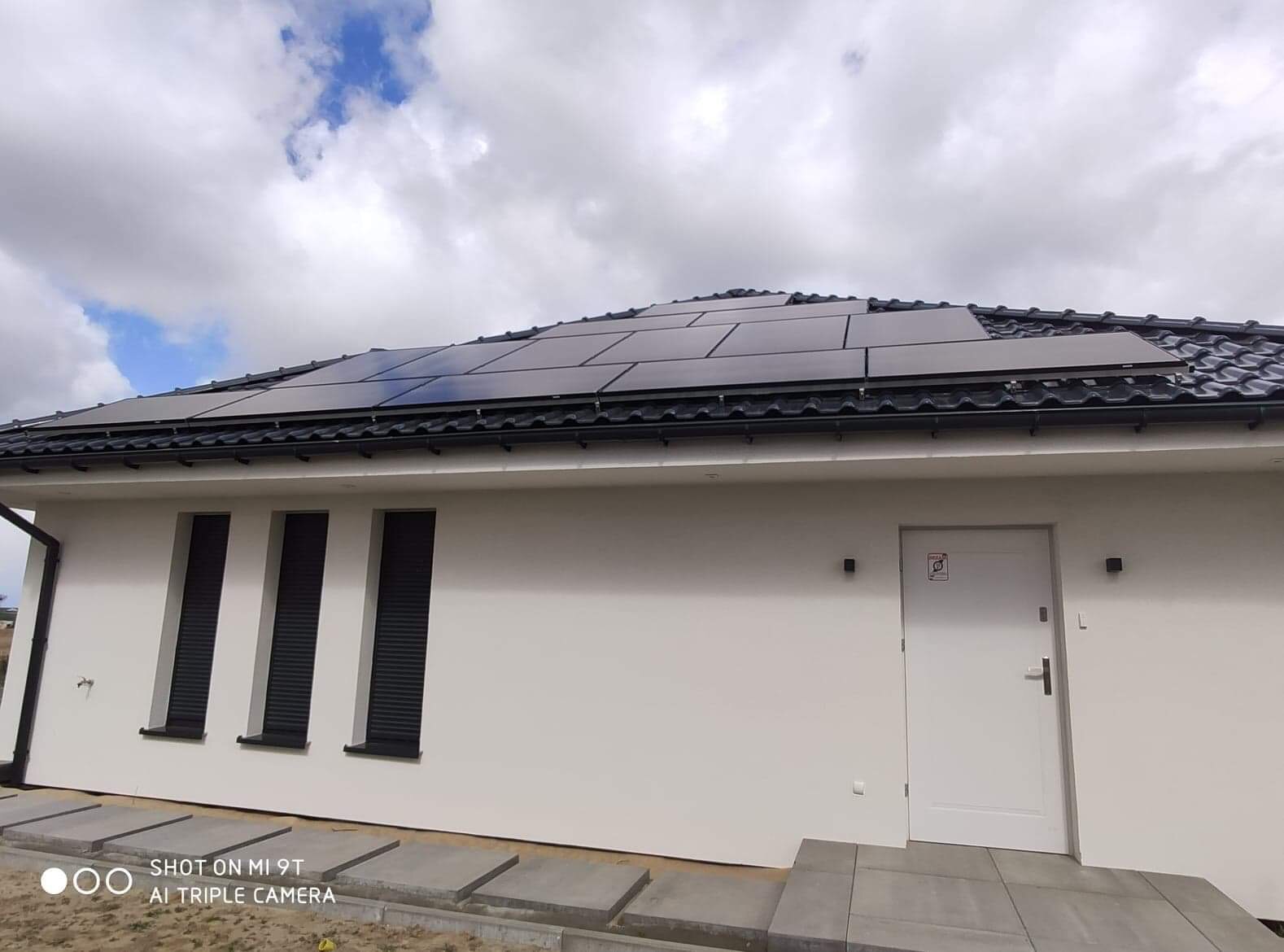 Panele słoneczne na dachu nowoczesnego, parterowego domu z trzema oknami z żaluzjami zewnętrznymi i białymi ścianami, widoczne betonowe płyty chodnikowe i fragment pochmurnego nieba.