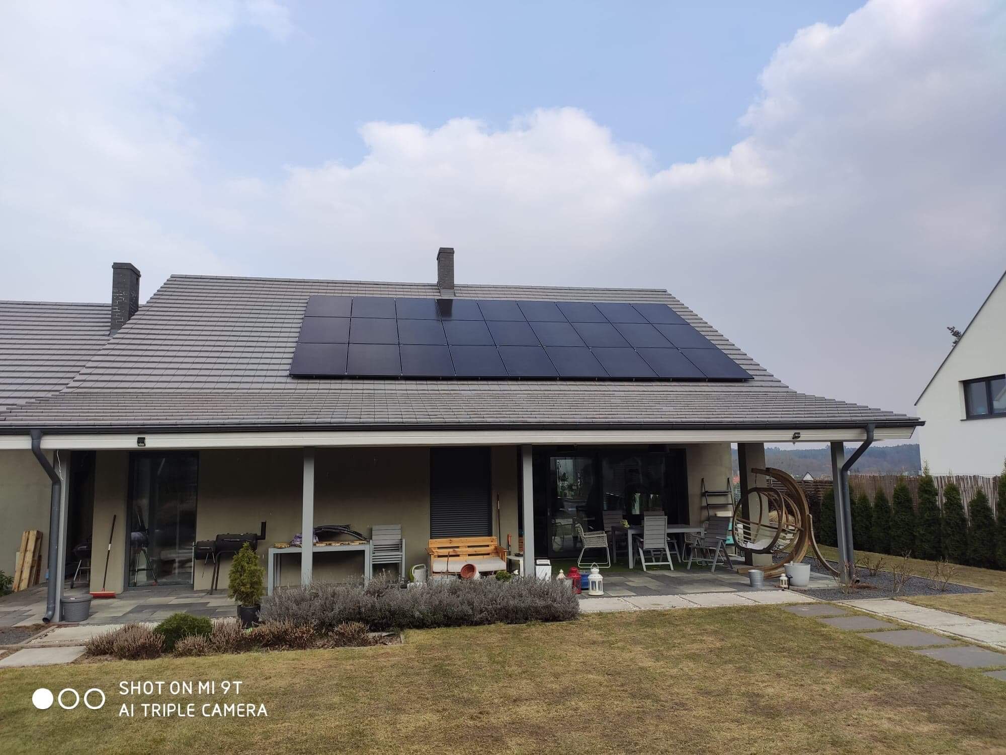 Dom jednorodzinny z szarym dachem i czarnymi panelami słonecznymi na połaci, widok z ogrodu w pochmurny dzień.