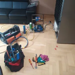 Sprzęt serwisowy rozłożony na parkiecie podczas naprawy lodówki: pompa próżniowa, butle z gazem, manometry, torby z narzędziami, śrubokręty i szmatki.