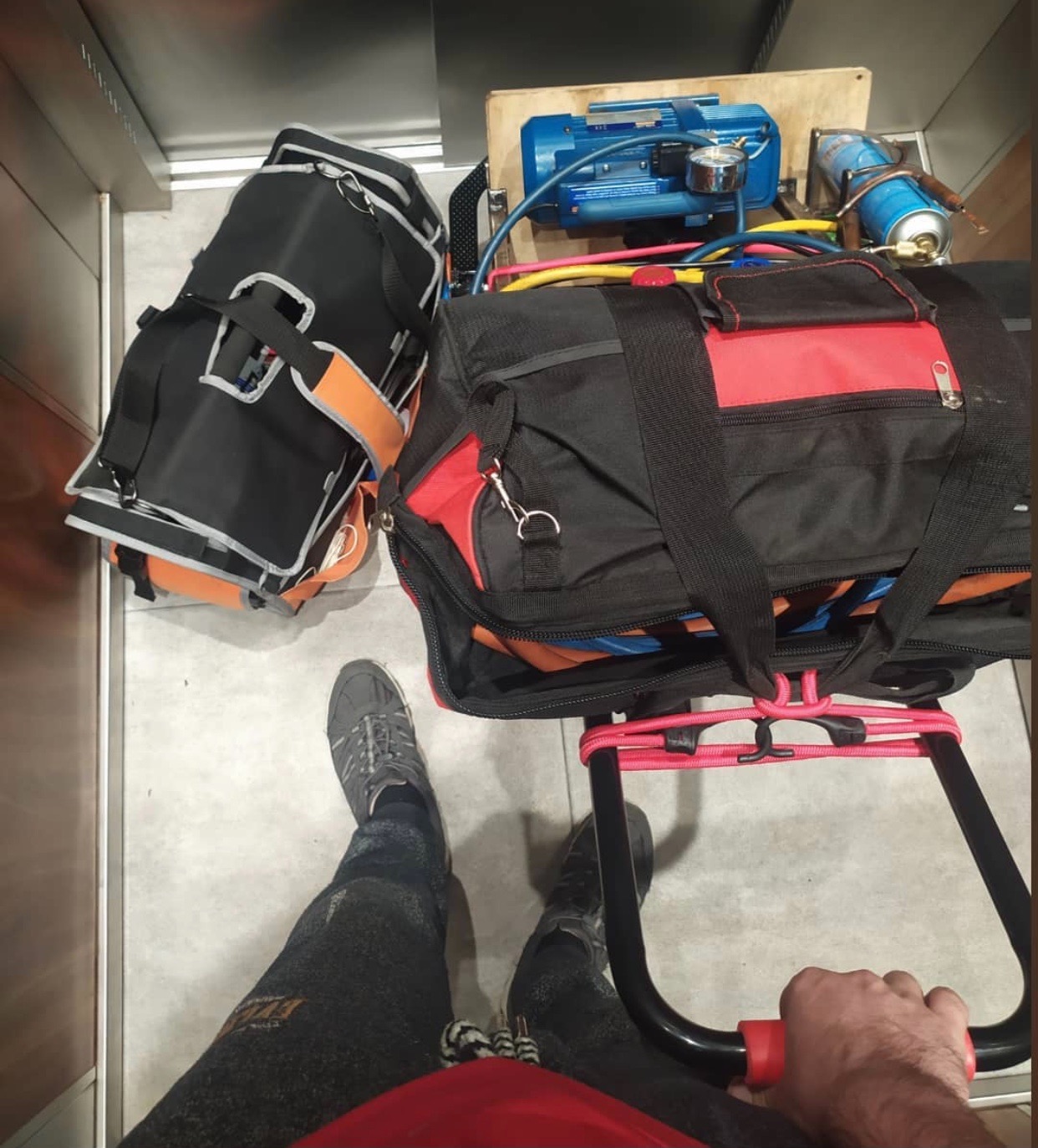 Technik w windzie transportuje sprzęt do naprawy lodówek: torby narzędziowe, butla z gazem, pompa próżniowa na wózku transportowym.