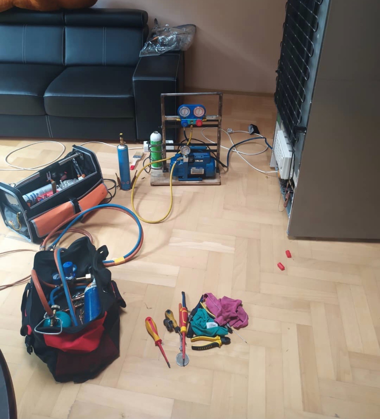 Sprzęt serwisowy rozłożony na parkiecie podczas naprawy lodówki: pompa próżniowa, butle z gazem, manometry, torby z narzędziami, śrubokręty i szmatki.