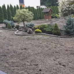 Przygotowany teren pod trawnik z obrzeżem z kostki wokół rabat z drzewami i krzewami ozdobnymi, studnia w tle.