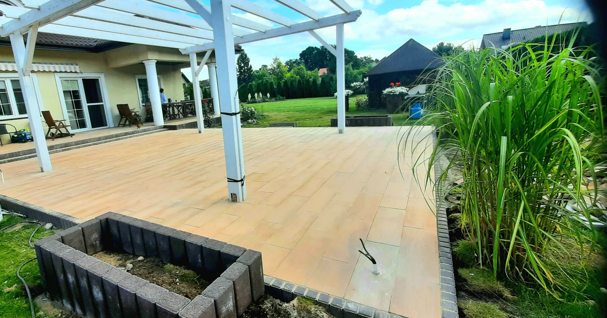 Jasna, nowa nawierzchnia z kostki brukowej na tarasie z białą pergolą, widok na zadbany ogród i domek ogrodowy w tle.