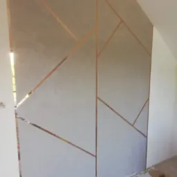 Ściana w pokoju po dekoracyjnym malowaniu z geometrycznym wzorem w kolorze miedzi, widoczne ślady farby na podłodze zabezpieczonej folią.