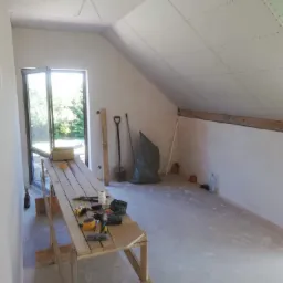 Poddasze w trakcie remontu, widoczne płyty kartonowo-gipsowe na skośnym suficie, rusztowanie z narzędziami, łopata, worek na śmieci i otwarte okno w tle.