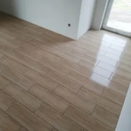 Ułożone pod kątem płytki podłogowe imitujące drewno, widoczne ślady czyszczenia w rogu pomieszczenia z oknem.