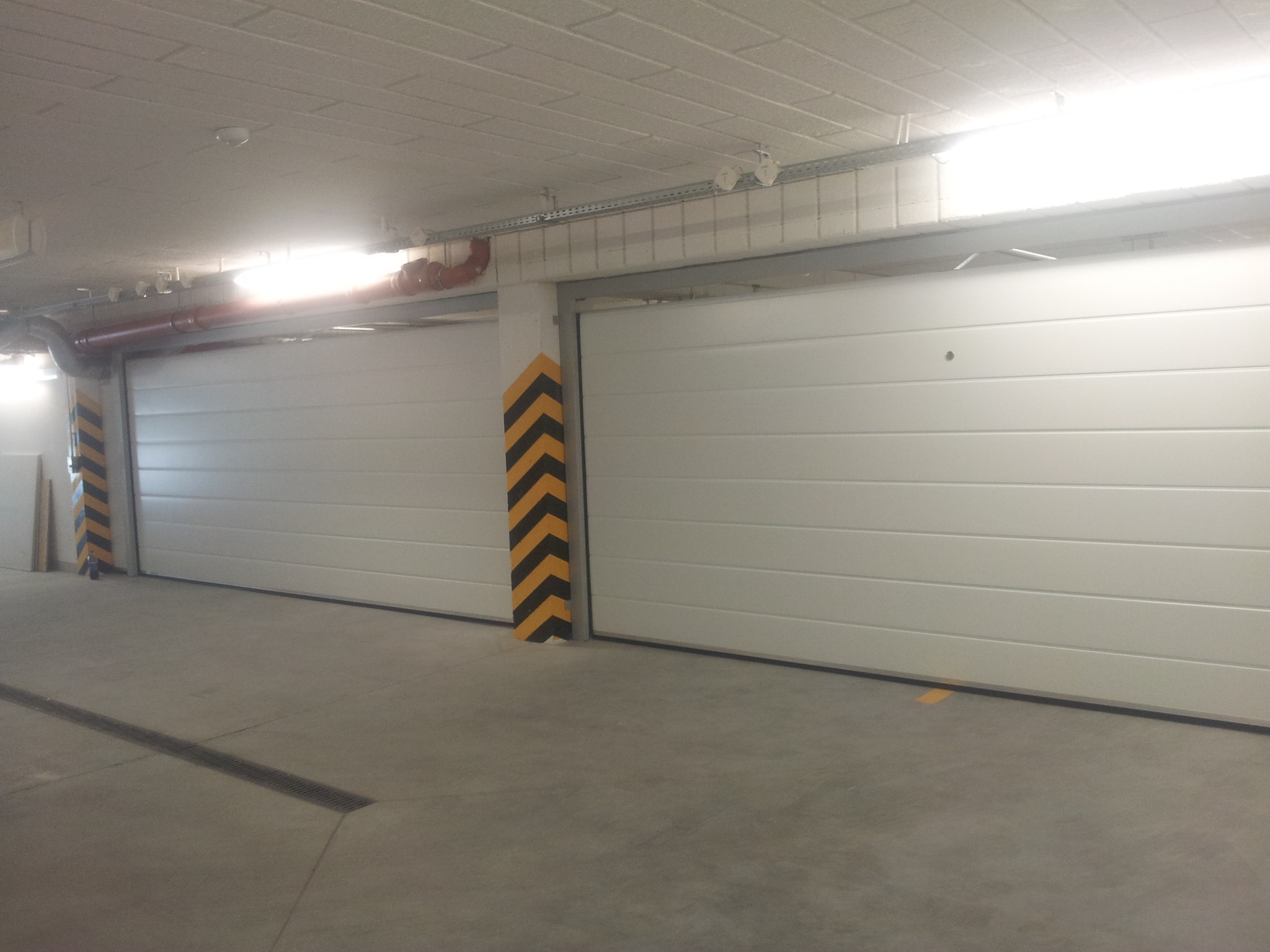 Wnętrze garażu podziemnego z dwiema białymi segmentowymi bramami, kolumny oznaczone czarno-żółtymi pasami ostrzegawczymi, widoczne instalacje pod sufitem.