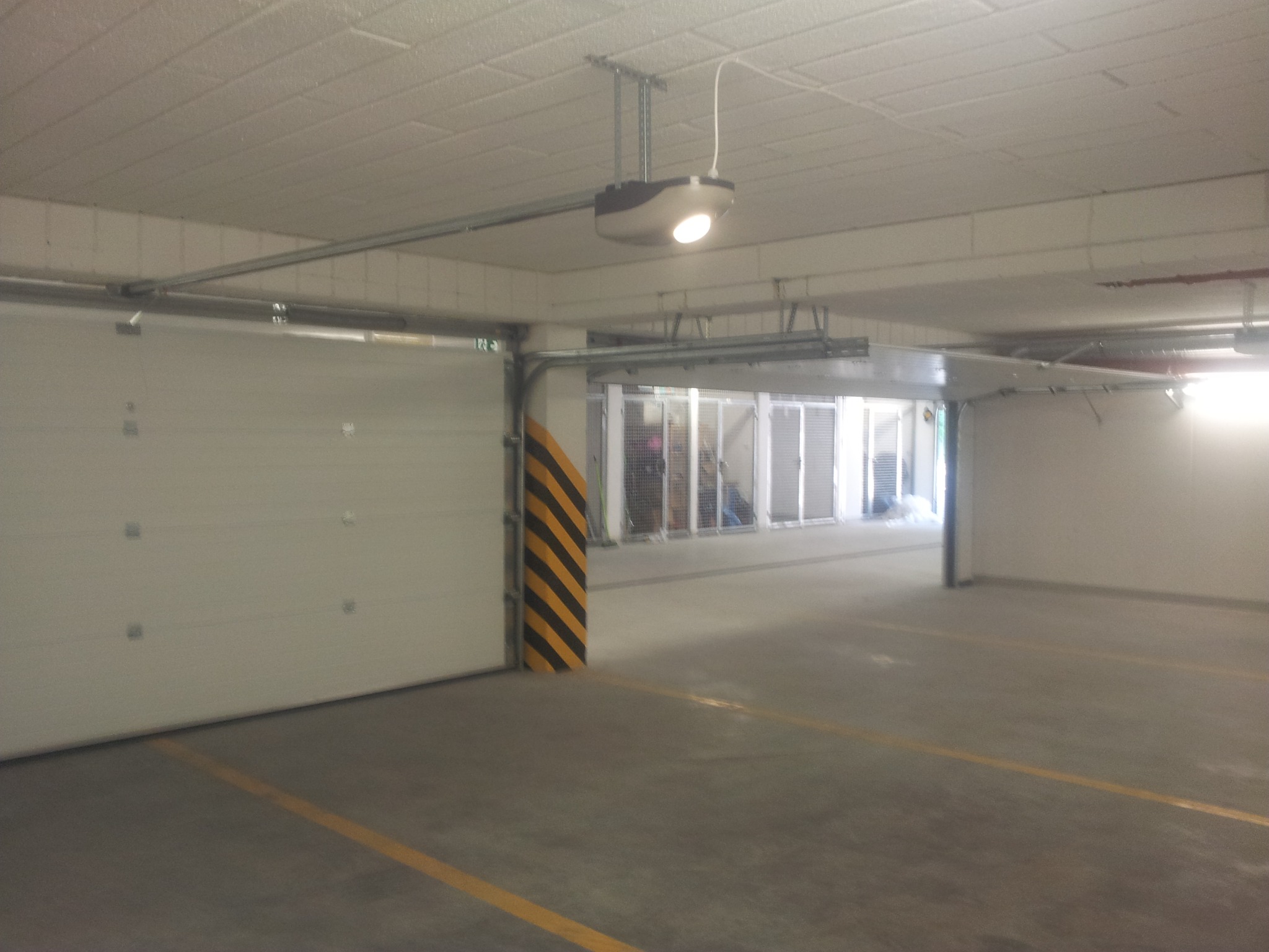 Otwarta segmentowa brama garażowa z automatycznym napędem zamontowanym pod sufitem w hali garażowej, widoczny fragment słupa z czarno-żółtymi pasami ostrzegawczymi.