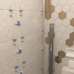 Układanie płytek w łazience z użyciem poziomicy i klipsów dystansowych, widoczne płytki heksagonalne w odcieniach bieli i złota oraz płytki z trójwymiarowym wzorem.