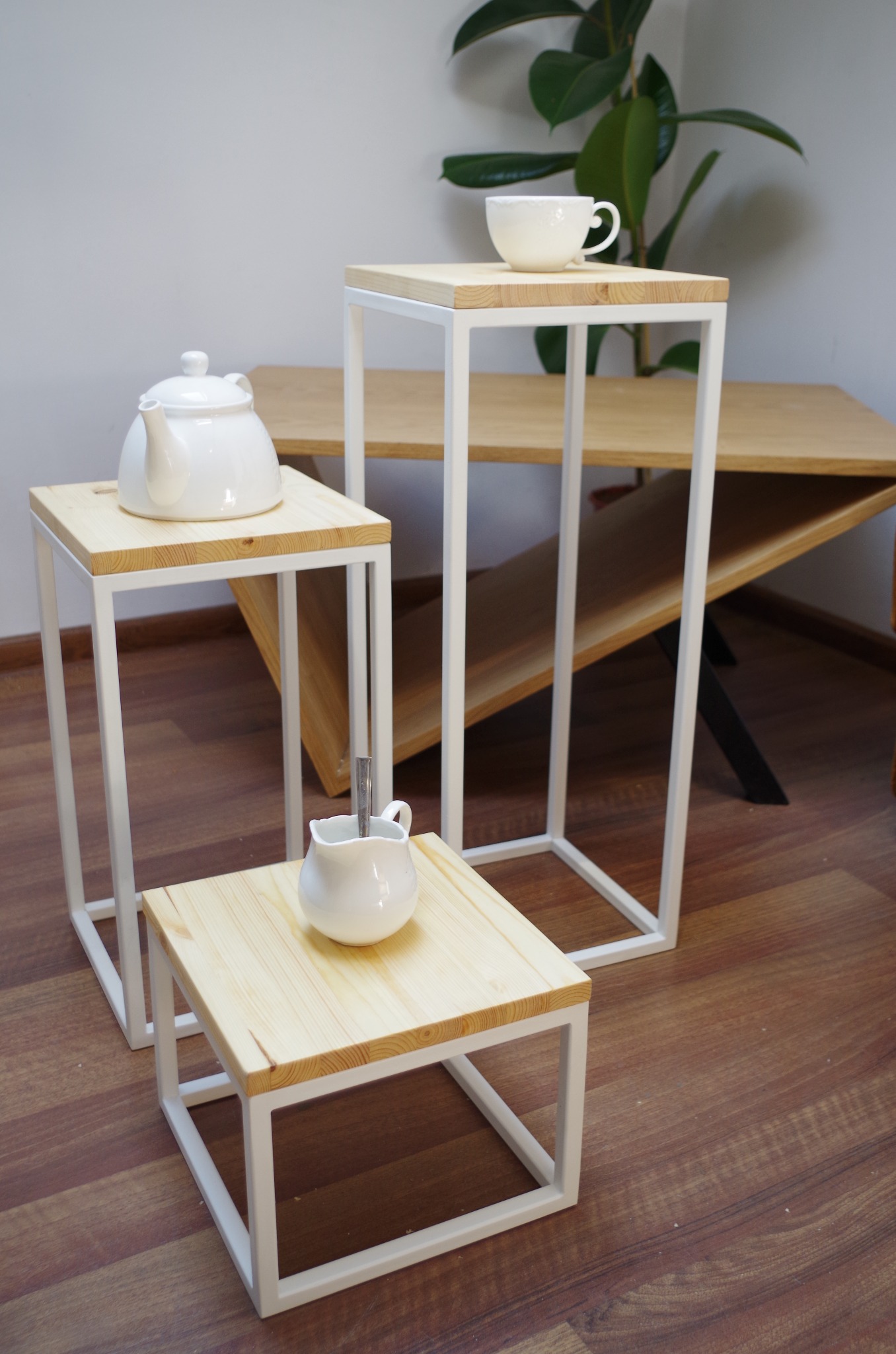 Trzy stoliki pomocnicze z drewnianymi blatami i białymi metalowymi ramami, na każdym z nich ustawiona biała porcelana: imbryk, dzbanuszek z łyżeczką i filiżanka, w tle drewniany stół i roślina...