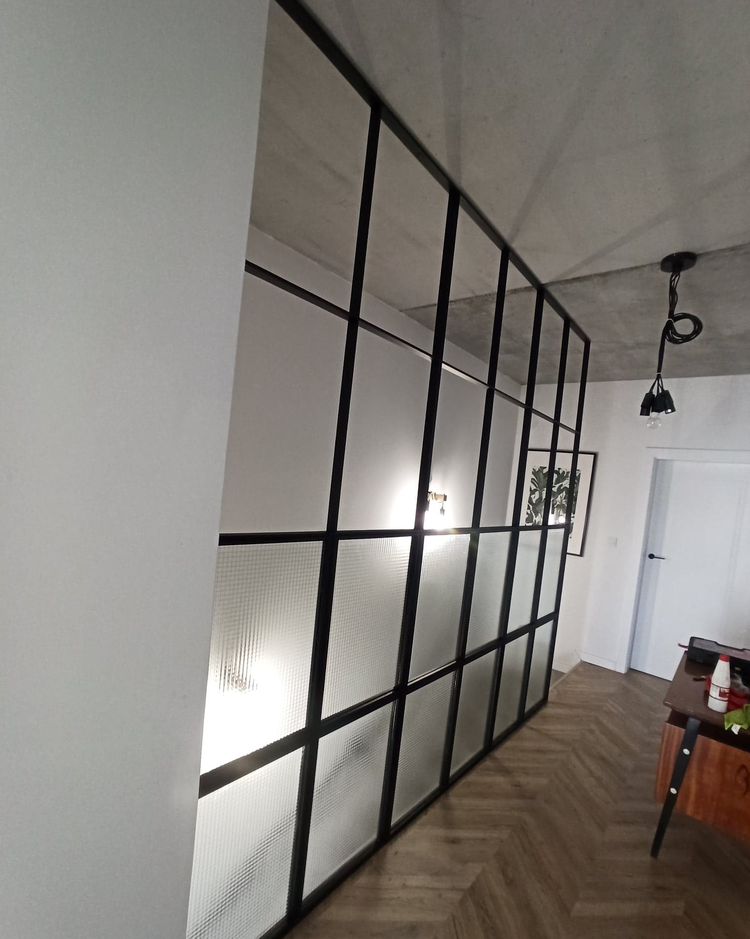 Metalowa ścianka działowa z matowym szkłem w czarnej ramie oddzielająca przestrzeń w nowoczesnym wnętrzu z parkietem w jodełkę i betonowym sufitem.