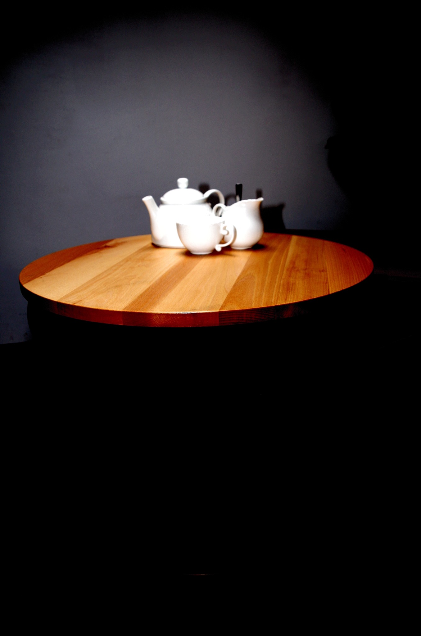Okrągły blat z jasnego drewna z zestawem do herbaty w białej porcelanie na tle ciemnoszarej ściany, oświetlony punktowo