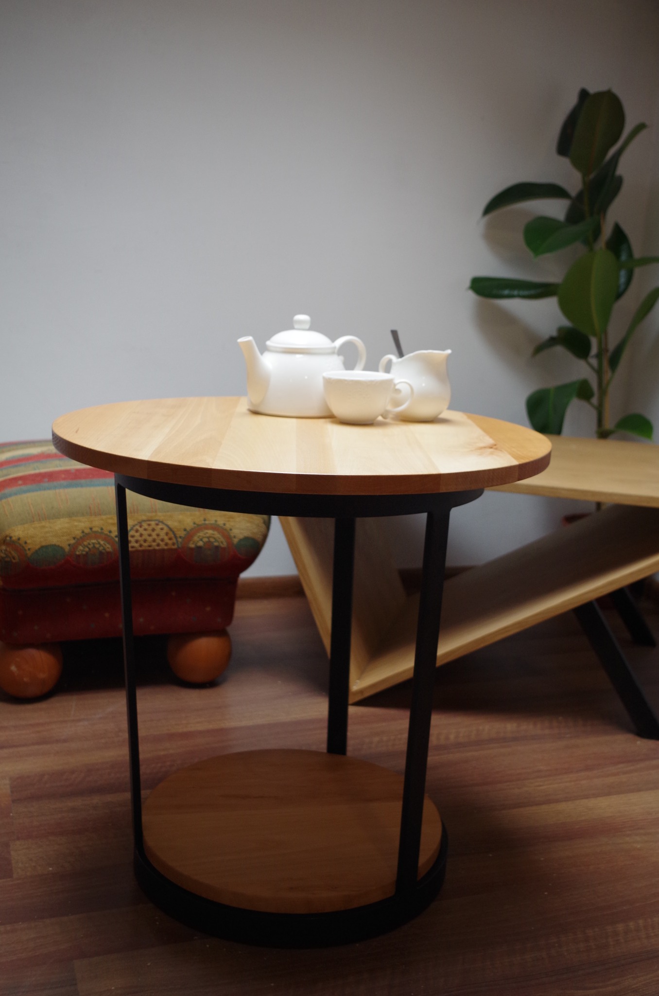 Okrągły stolik kawowy z blatem z jasnego drewna i czarnymi metalowymi nogami, na nim biały serwis do herbaty, w tle pufa w paski i roślina doniczkowa.