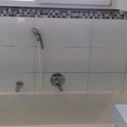 Wnętrze łazienki z wanną, armaturą i białymi płytkami, dekoracyjne mozaiki w górnej części ściany, woda leje się z prysznica.