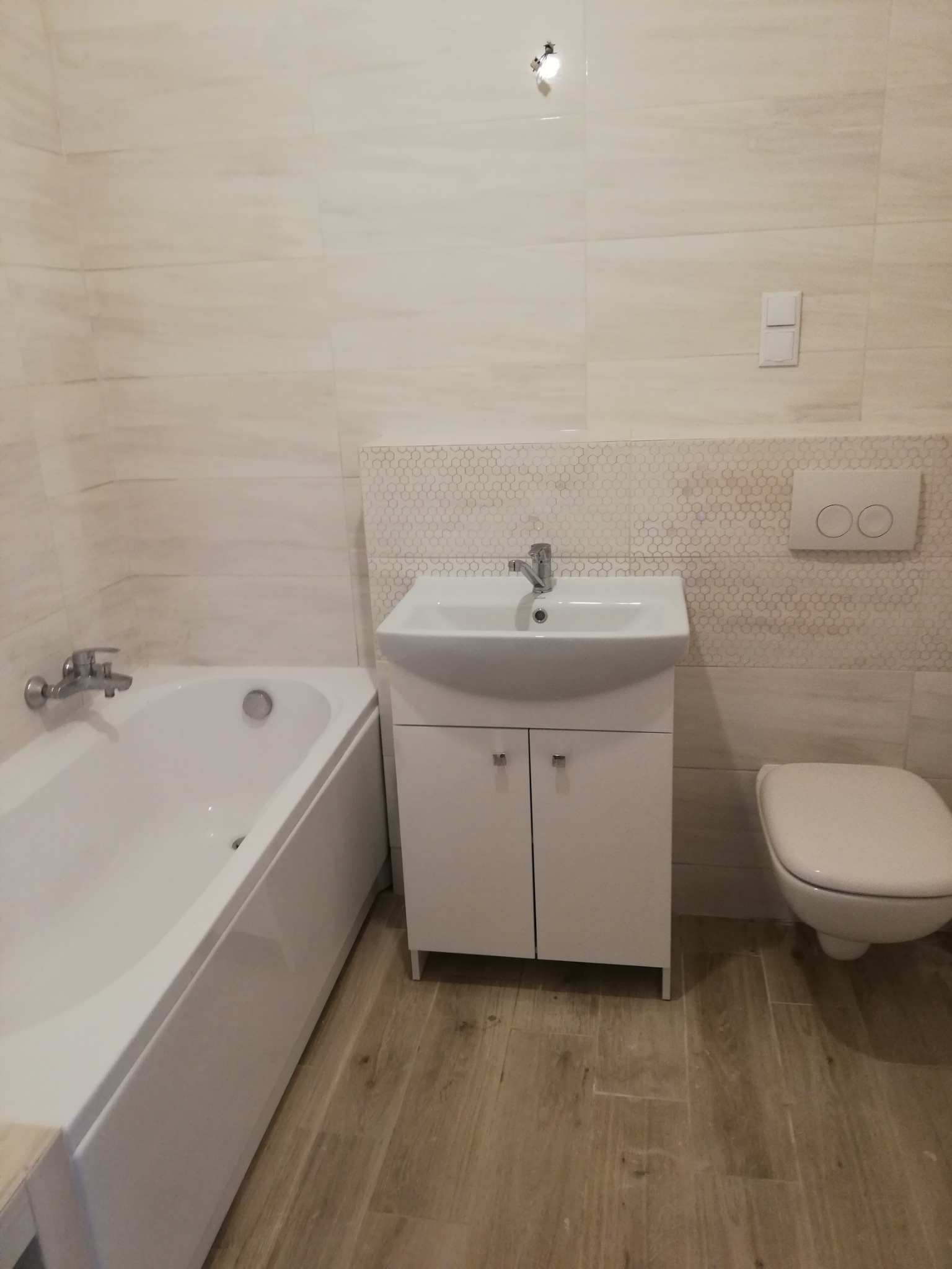 Jasna łazienka po remoncie: wanna, umywalka z szafką, toaleta i płytki imitujące drewno na podłodze oraz beżowe płytki na ścianach z dekoracyjnym pasem w kształcie plastra miodu.