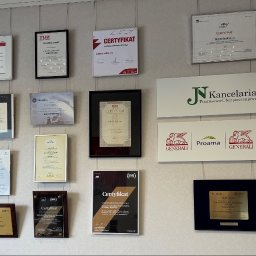KANCELARIA FINANSOWO-UBEZPIECZENIOWA JN SPÓŁKA Z OGRANICZONĄ ODPOWIEDZIALNOŚCIĄ - Ściana z licznymi oprawionymi certyfikatami i dyplomami dla Violetty Józefowskiej, tablicami 'JN Kancelaria Finansowo-Ubezpieczeniowa' i logotypami ubezpieczycieli.