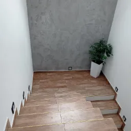 Klatka schodowa z drewnopodobnymi płytkami na stopniach i szarą ścianą dekoracyjną, oświetlenie punktowe w ścianie, roślina doniczkowa w białej osłonce.