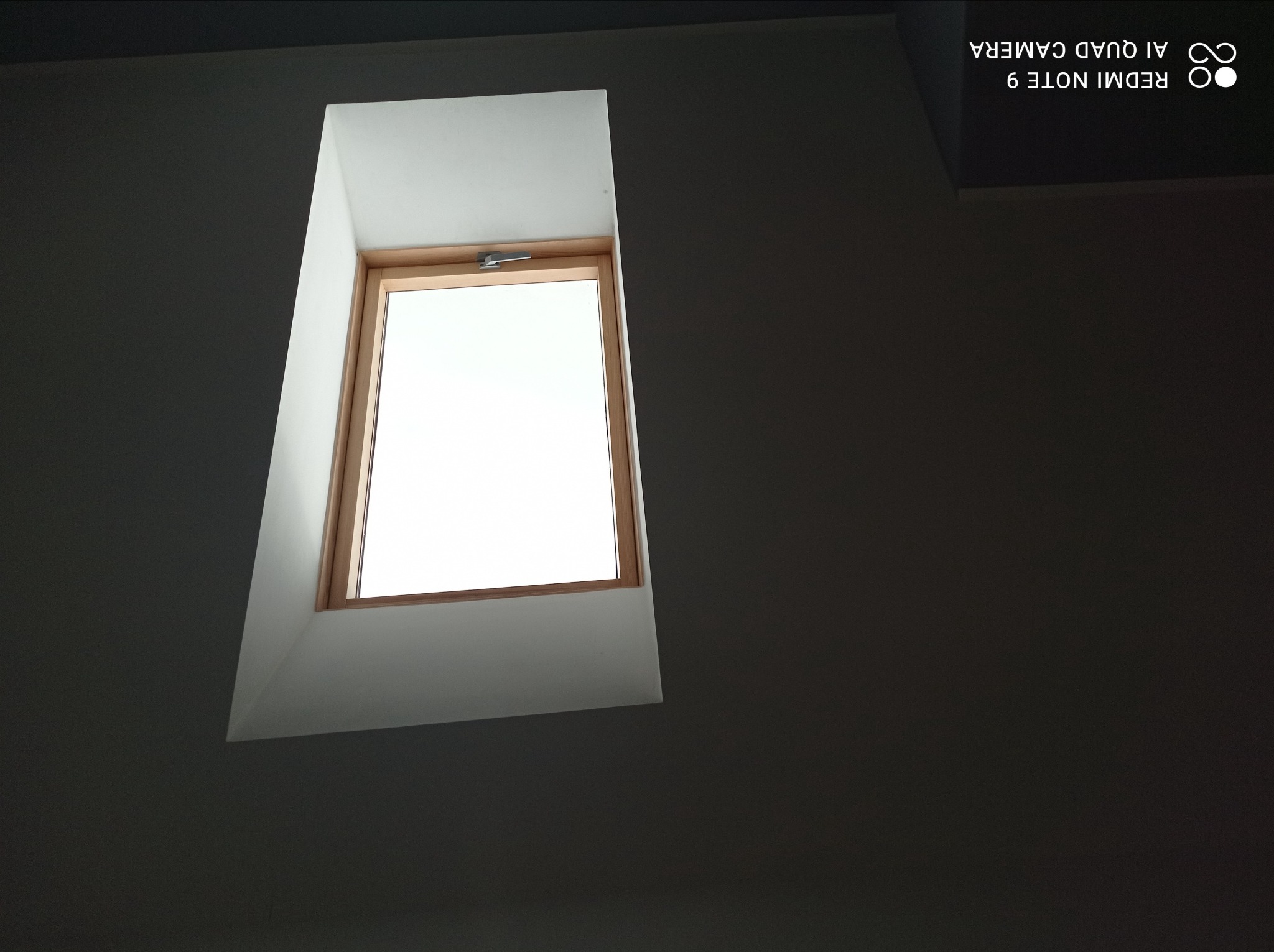 Okno dachowe z drewnianą ramą, osadzone w białej wnęce w szarej ścianie pod skośnym sufitem. Widoczny mechanizm otwierania okna.