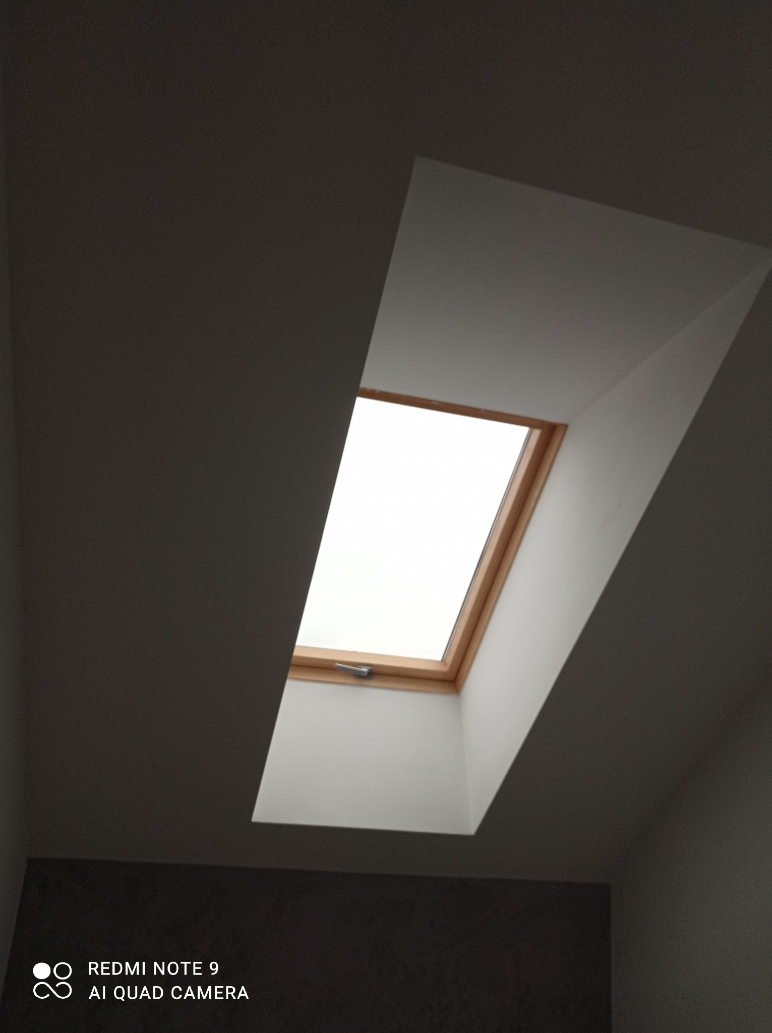 Okno dachowe z drewnianą ramą, zamontowane w skośnym suficie, widok od dołu, z widocznym uchwytem do otwierania.