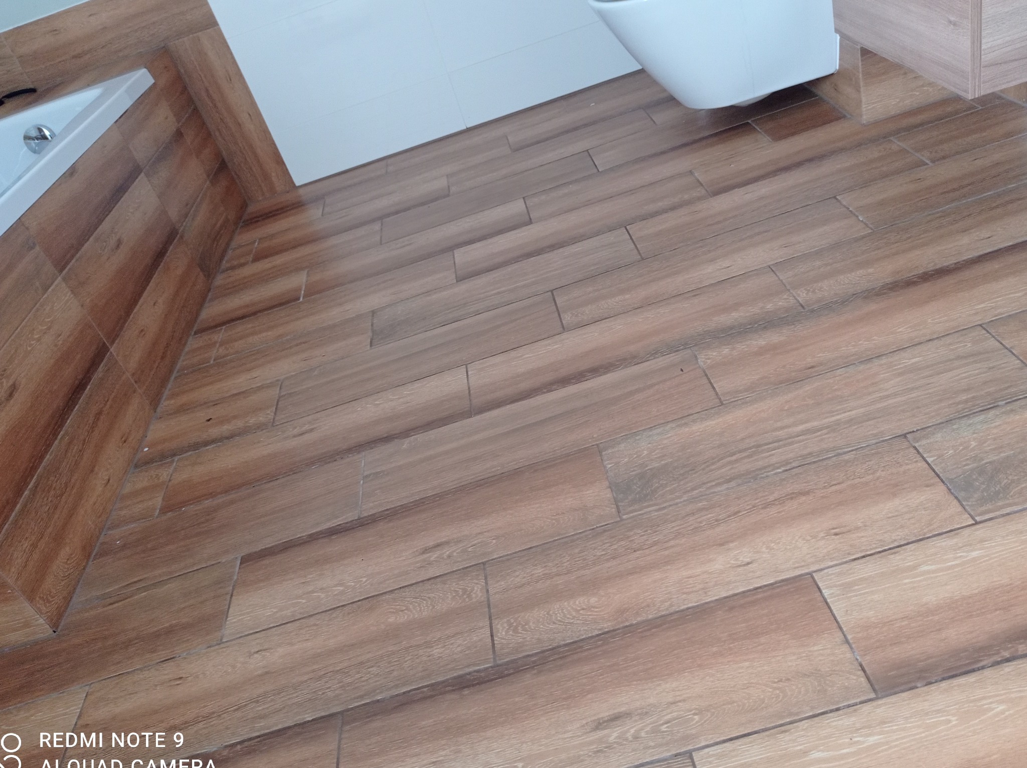 Ułożona pod kątem brązowa podłoga z płytek imitujących drewno w łazience, widoczny fragment toalety i obudowy wanny.