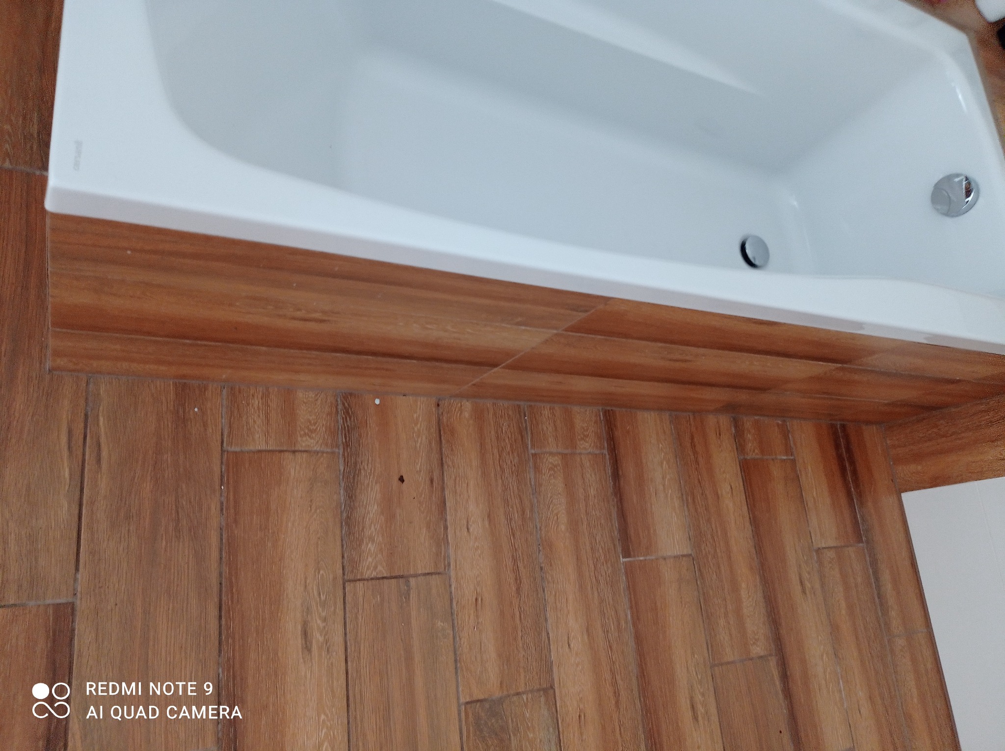 Biała wanna obudowana płytkami imitującymi drewno, z widocznym odpływem i przelewem, oraz podłoga wyłożona identycznymi płytkami, tworząca spójną aranżację łazienki.