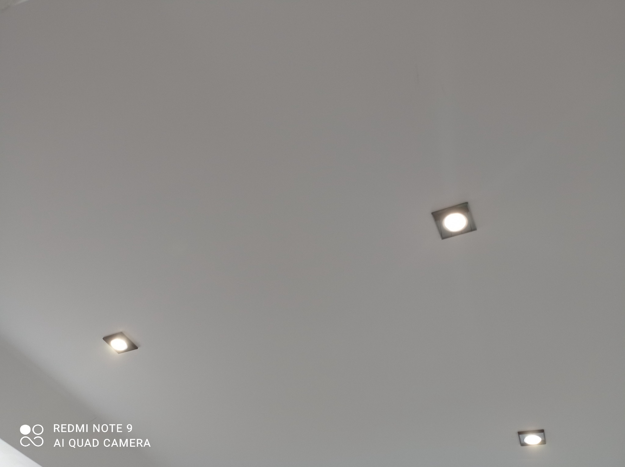 Biały sufit z trzema kwadratowymi oprawami oświetleniowymi LED, rozmieszczonymi w rogach kadru, widoczne delikatne smugi na powierzchni.
