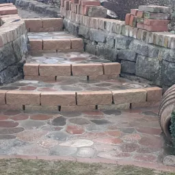 Kamienne schody ogrodowe z brukowaną ścieżką, otoczone niskim murkiem z kamienia i cegły, z dekoracyjną donicą z rośliną obok.