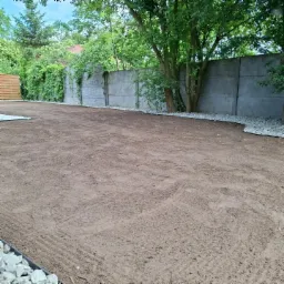 Przygotowany teren pod trawnik z widocznymi śladami po bronowaniu, obrzeżami z białego kamienia i betonowym ogrodzeniem porośniętym bluszczem w tle.