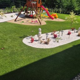 Usługi Ogrodnicze - Mieniący się zielony trawnik z placem zabaw dla dzieci, piaskownicą i ozdobną wyspą z białych kamieni, roślinami i figurką. W tle żywopłot i ogrodzenie.