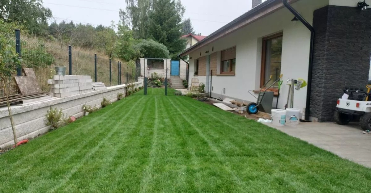Świeżo skoszony trawnik z wyraźnymi pasami, graniczący z murem oporowym z betonowych bloków i młodymi drzewkami, w tle dom z elementami wykończenia z ciemnego kamienia i zabawkowy pojazd.