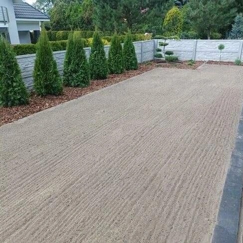 Przygotowany teren pod trawnik z widocznymi śladami po bronowaniu, obok rząd tuj i dekoracyjny mur z betonu architektonicznego.