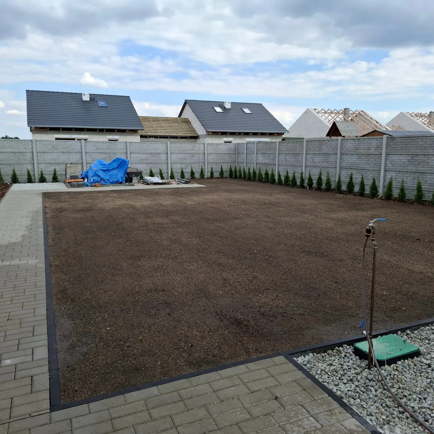 Przygotowany teren pod trawnik, ziemia wyrównana i nawodniona, widoczny system nawadniania, obrzeża z kostki brukowej, w tle ogrodzenie z prefabrykatów betonowych i nowe domy.