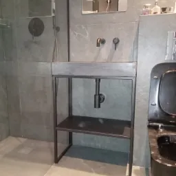 Nowoczesna łazienka w Lublinie z szarą armaturą i umywalką na metalowym stelażu, widoczny fragment kabiny prysznicowej i toalety w ciemnym kolorze.