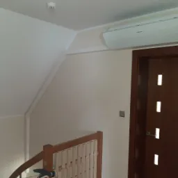 Biała jednostka klimatyzacyjna Samsung zamontowana wysoko na ścianie w pokoju na poddaszu, obok brązowych drzwi z przeszkleniami, widoczna kratka wentylacyjna na suficie.