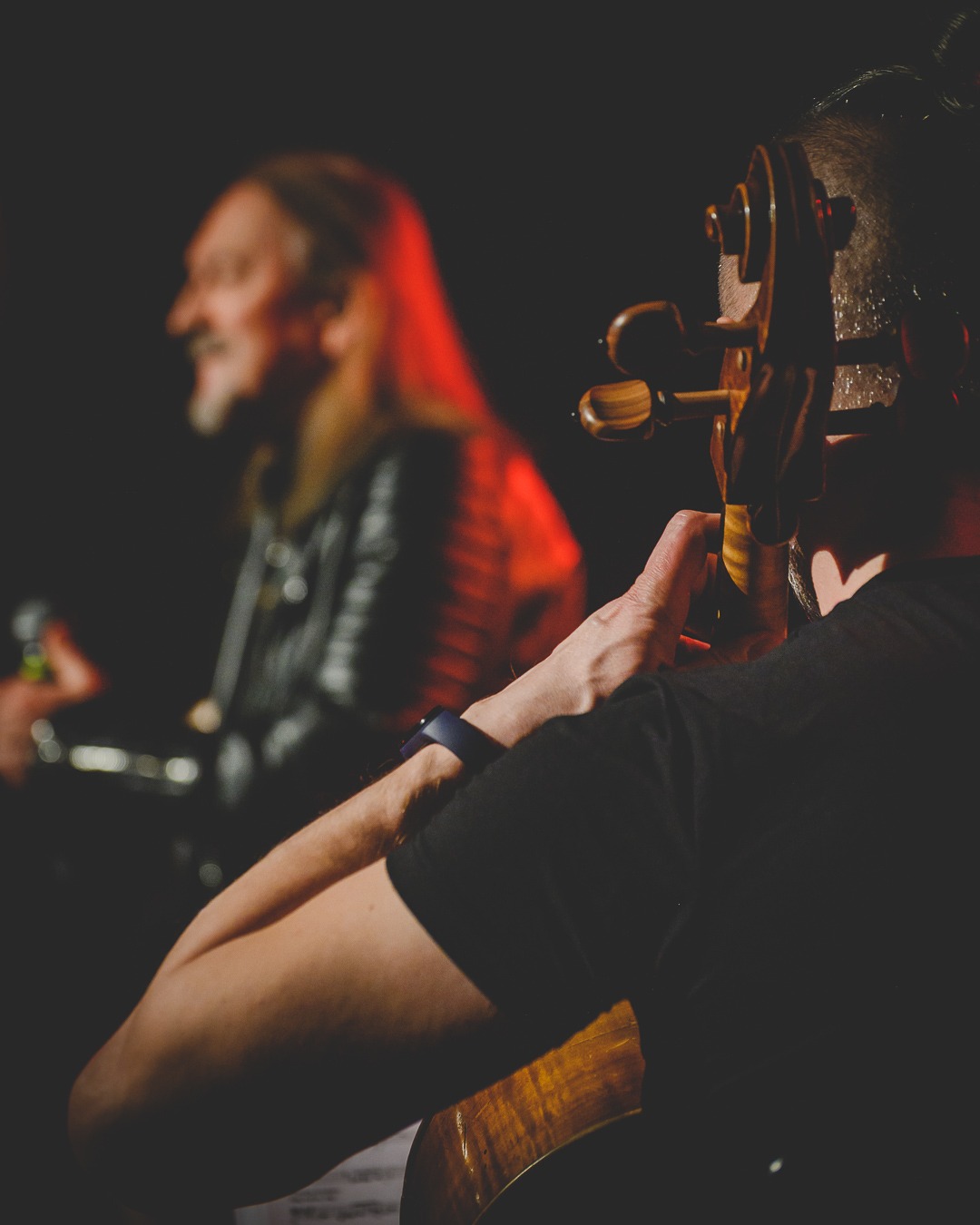 Muzyk grający na kontrabasie na scenie w trakcie koncertu, z widocznym drugim muzykiem w tle oświetlonym czerwonym światłem.
