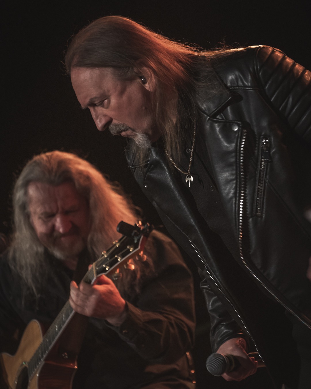 Fotografia koncertowa z Goleniowa: wokalista w skórzanej kurtce i gitarzysta z akustyczną gitarą podczas występu na scenie w ciemnym oświetleniu.