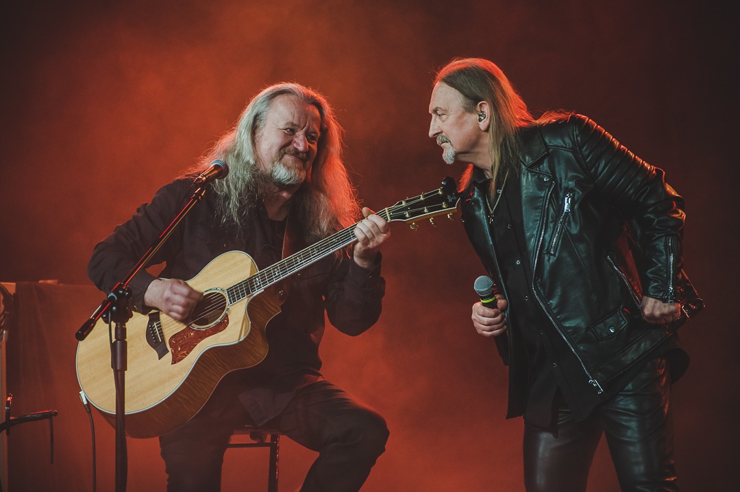 Dwóch muzyków rockowych występuje na scenie: jeden gra na gitarze akustycznej siedząc na krześle, drugi śpiewa do mikrofonu, obaj ubrani w czarne stroje na tle czerwonego oświetlenia.