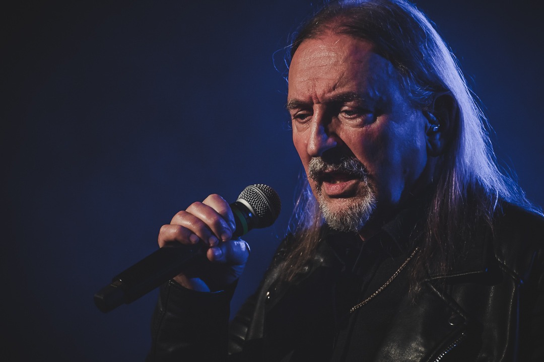 Mężczyzna z długimi włosami i brodą, w skórzanej kurtce, śpiewa do mikrofonu na ciemnym tle, oświetlony niebieskim światłem.