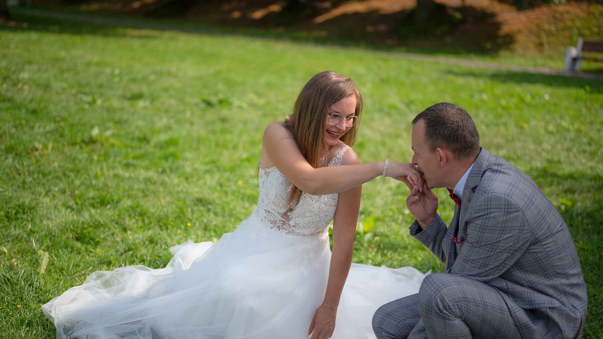 Panna młoda w okularach i koronkowej sukni siedzi na trawie, a pan młody w szarej kraciastej marynarce całuje ją w dłoń podczas sesji ślubnej.