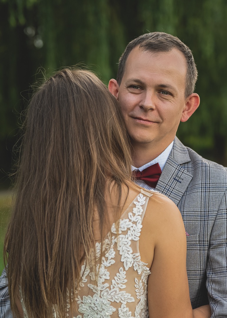 Uśmiechnięty mężczyzna w szarej marynarce w kratę i bordowej muszce obejmuje kobietę w sukni z koronkowymi aplikacjami na plecach, widok z tyłu.