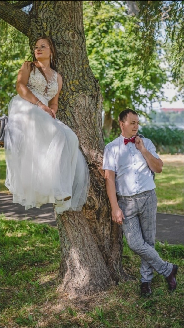 Panna młoda w koronkowej sukni siedzi na gałęzi drzewa, obok stoi pan młody w szarej kraciastej marynarce i bordowej muszce.