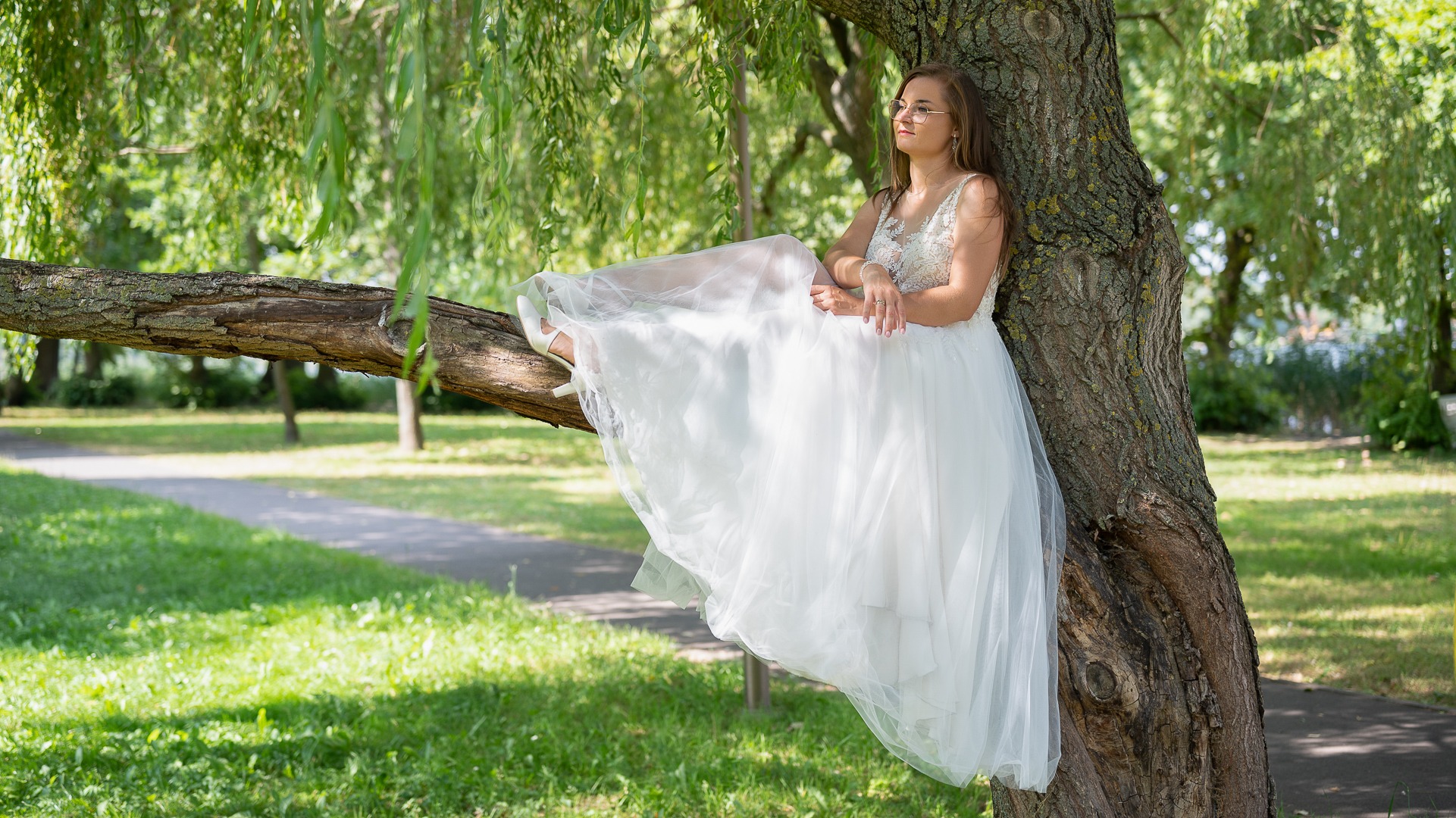 Kobieta w białej sukni ślubnej siedzi na gałęzi drzewa w parku, spoglądając w dal.