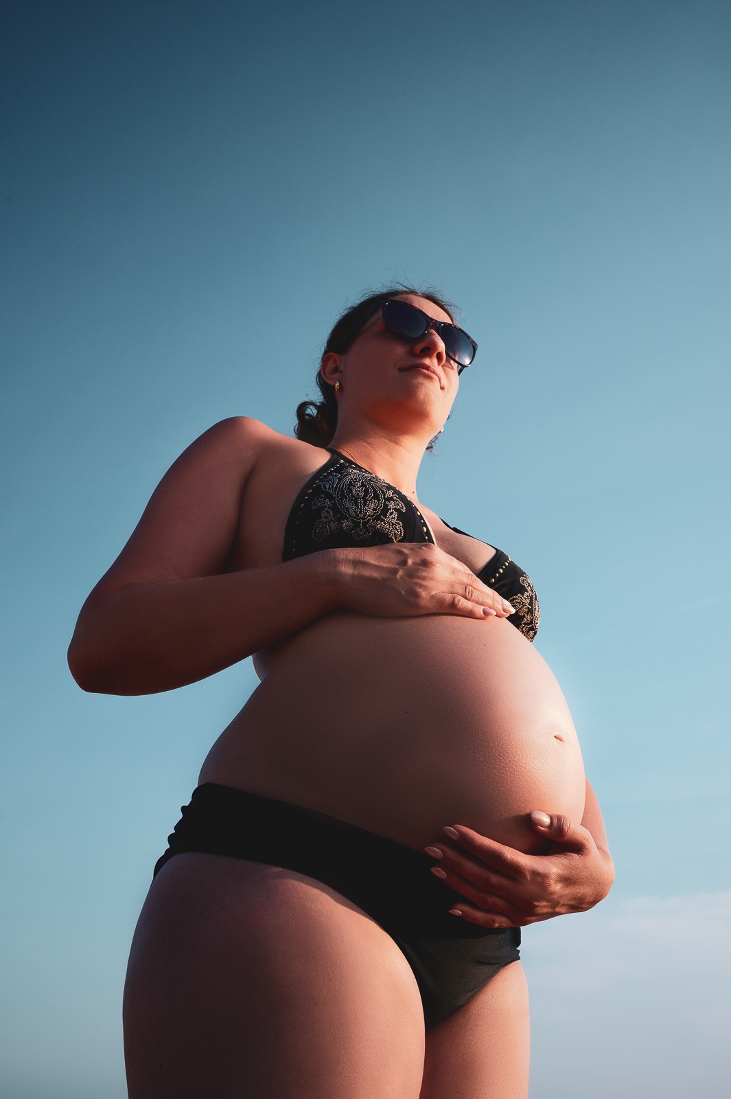 Kobieta w bikini i okularach przeciwsłonecznych, trzymająca ręce na ciążowym brzuchu, sfotografowana z dołu na tle błękitnego nieba.
