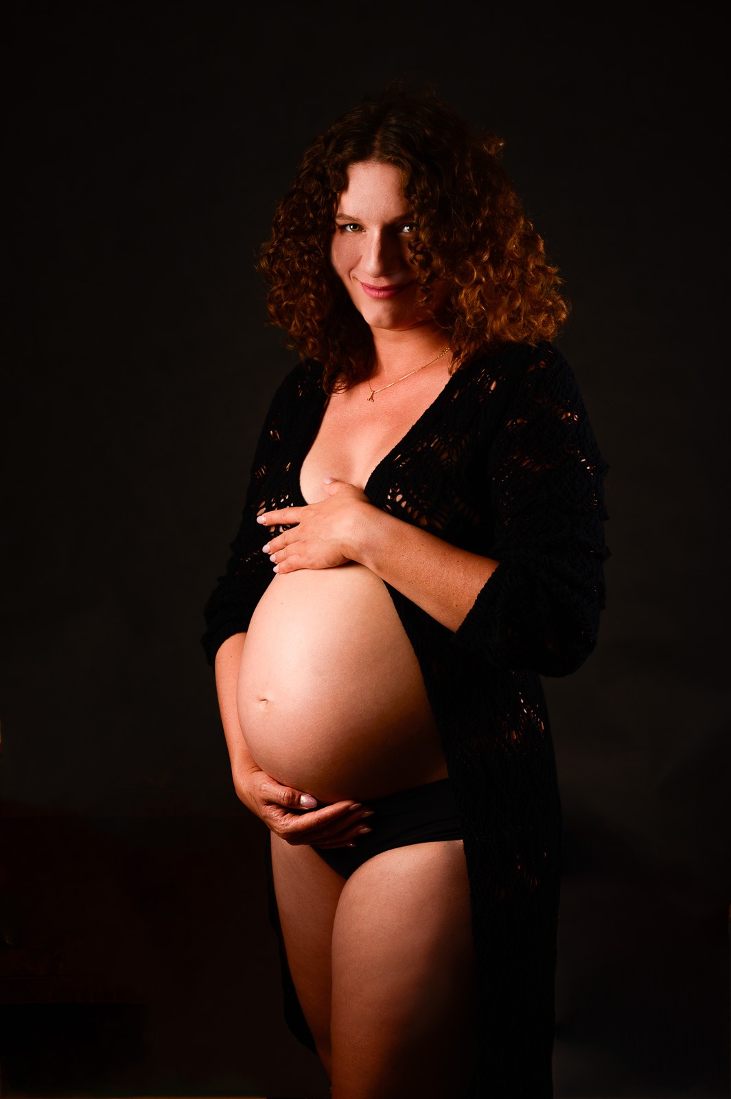 Kobieta w zaawansowanej ciąży, z kręconymi włosami i delikatnym naszyjnikiem, pozuje w czarnej, ażurowej narzutce na ciemnym tle, dłonie ułożone na brzuchu.